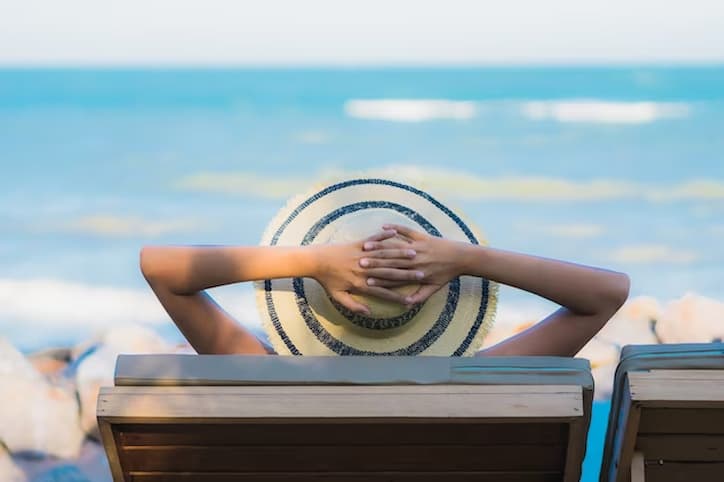 Mulher deitada em uma cadeira na praia com as mãoas na cabeça aproveitando os feriados de 2024