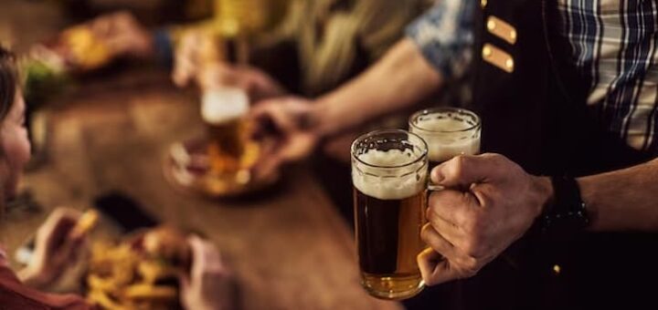 Close na mão de um garçom segurando duas canecas de cervejas enquanto um grupo de maigos comemoram em um bar em Moema.